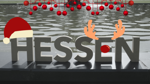 Buchstaben stehen vor einem Brunnen. Sie ergeben das Wort hessen. Das H trägt eine Nikolausmützem das E ein Rentiergeweih mit roter Nase.