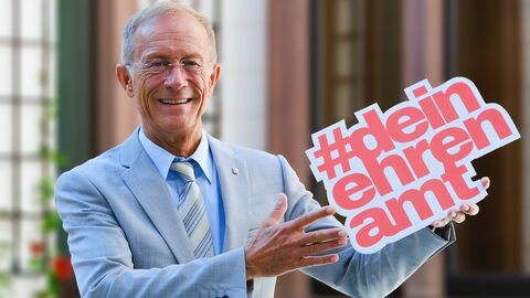Staatsminister Wintermeyer hält das Logo #deinehrenamt hoch und lächelt in die Kamera.