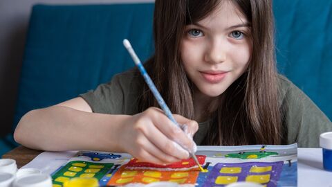 Junges Kind sitzt am Tisch und malt mit Wasserfarben. Es guckt in die Kamera