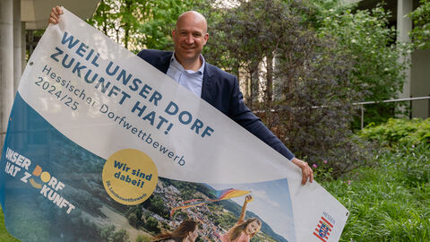 Heimatminister Jung steht vor Bäumen und halt ein Plakat "Weil unser Dorf Zukunft hat - Hessischer Dorfwettbewerb 2024/25" hoch.