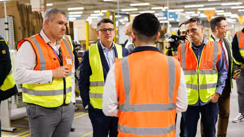 Ministerpräsident Rhein steht mit weiteren Personen in einer Lagerhalle. Sie tragen alle Warnwesten und sind ins Gespräch vertieft.