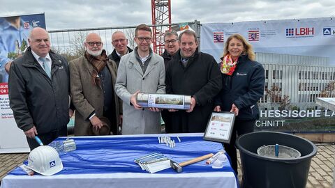 7 Personen stehen nebeneinander. Zwei davon halten eine Zeitkapsel hoch. Sie stehen draußen auf einem Baugelände mit Bauzaun im Hintergrund.