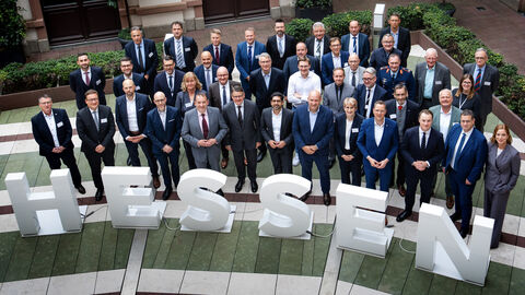 Über 30 Personen stehen in 3 Reihen nebeneinander in einem Innenhof und schauen nach oben in die Kamera. Vor ihnen sind 1 Meter hohe Buchstaben platziert, die das Wort "Hessen" ergeben.