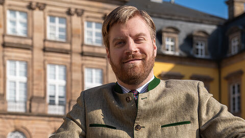 Ein Mann lächelt in die Kamera. Er sitzt draußen und hinter ihm ist ein Gebäude aus Stein.