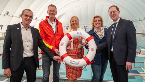 V.l.n.r.: Matthias Wörner (Vorsitzender Erster Offenbacher Schwimmclub von 1896 e.V.), Michael Hohmann (Präsident Deutsche Lebens-Rettungs-Gesellschaft Landesverband Hessen e.V. (DLRG)), Sportministerin Diana Stolz, Juliane Kuhlmann (Präsidentin Landessportbund Hessen (lsb h)) und Michael Scragg (Präsident Hessischer Schwimm-Verband e.V.) stehen in einer Schwimmhalle nebeneinander. Sportministerin Stolz hält einen Rettungsring vor sich und alle lächeln.